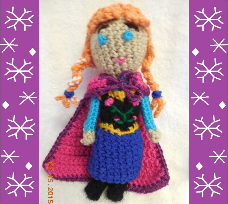 May include: Crocheted doll with red, purple, and blue clothing. The doll has orange hair in braids and blue eyes. The doll is wearing a cape and a dress with a floral design.