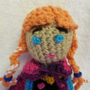 May include: A crocheted doll with orange hair, blue eyes, and a pink, purple, and blue dress. The doll is wearing a purple ribbon around its neck. The doll is sitting on a white surface. 03.15.2015