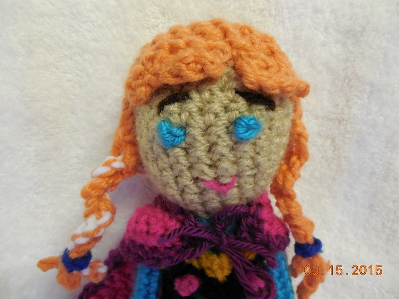 May include: A crocheted doll with orange hair, blue eyes, and a pink, purple, and blue dress. The doll is wearing a purple ribbon around its neck. The doll is sitting on a white surface. 03.15.2015