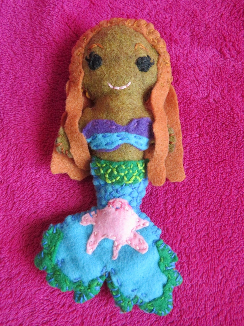 May include: A felt mermaid doll with long orange hair, a purple and blue top, a green and blue tail, and a pink starfish on the tail.