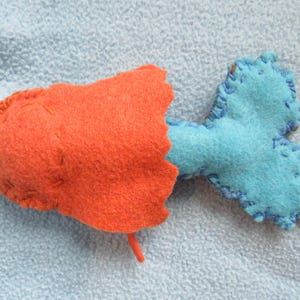 May include: A handmade felt fish toy with an orange body and a blue tail. The fish has a simple, stitched design.