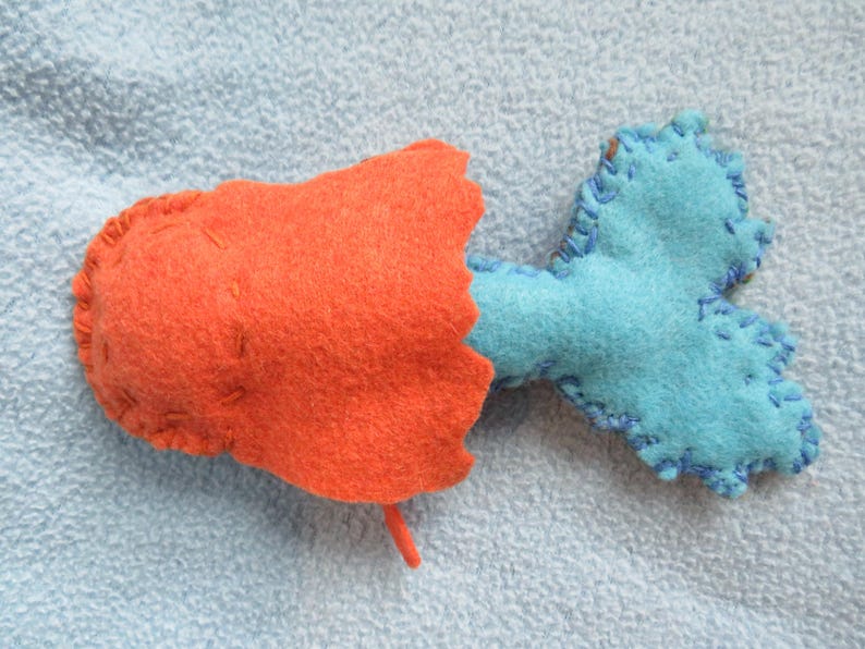 May include: A handmade felt fish toy with an orange body and a blue tail. The fish has a simple, stitched design.