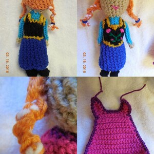 May include: A crocheted doll with long orange hair, wearing a blue dress with a black vest and a floral design. The doll is standing on a white surface.