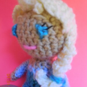 Op de afbeelding: Een gehaakte pop met een witblond pruikje, blauwe ogen en een roze mond. De pop draagt een blauwe jurk met witte accenten.