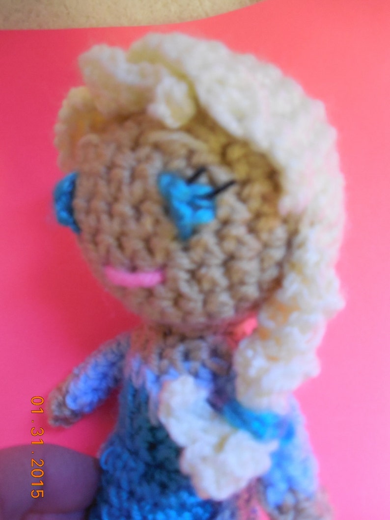 Op de afbeelding: Een gehaakte pop met een witblond pruikje, blauwe ogen en een roze mond. De pop draagt een blauwe jurk met witte accenten.