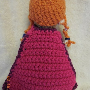 May include: A crocheted doll with a pink cape and orange hair. The doll is wearing a purple and pink crocheted dress.