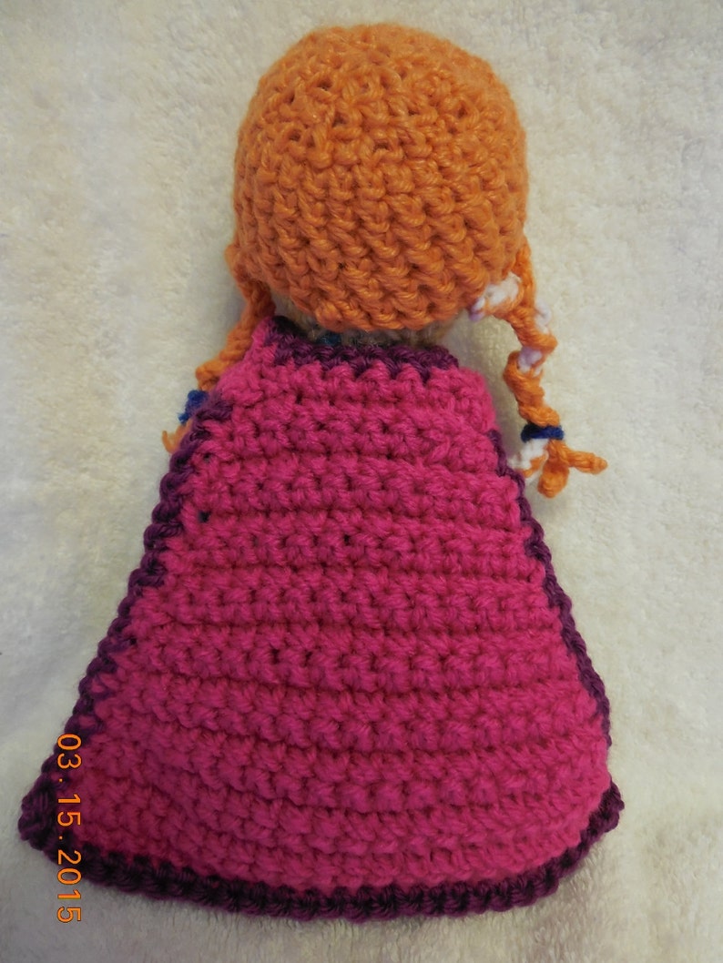 May include: A crocheted doll with a pink cape and orange hair. The doll is wearing a purple and pink crocheted dress.
