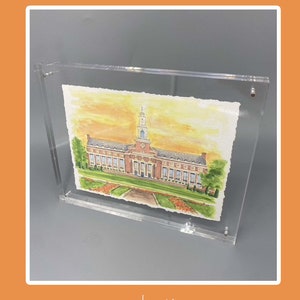 Pode incluir: Uma pintura em aquarela de um grande edif&iacute;cio de tijolos com uma torre central e um gramado verde, inserida em uma moldura de acr&iacute;lico transparente. A pintura apresenta tons quentes de c&eacute;u amarelo e laranja, &aacute;rvores verdes e um caminho.