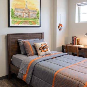 Pode incluir: Uma cama de solteiro com estrutura de madeira escura, apresentando um edredom cinza com listras laranja e padr&otilde;es geom&eacute;tricos. Uma almofada quadrada exibe o logotipo da Oklahoma State University. Acima da cama, h&aacute; uma aquarela emoldurada de um pr&eacute;dio.