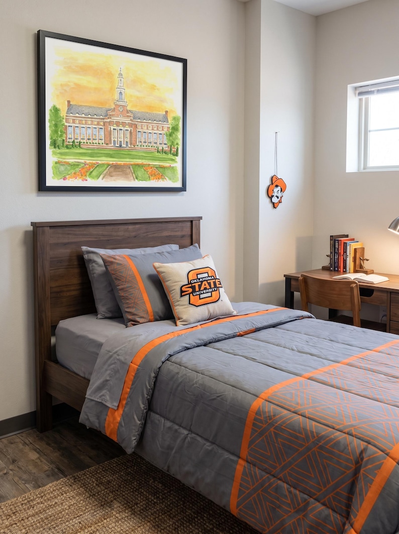 Pode incluir: Uma cama de solteiro com estrutura de madeira escura, apresentando um edredom cinza com listras laranja e padr&otilde;es geom&eacute;tricos. Uma almofada quadrada exibe o logotipo da Oklahoma State University. Acima da cama, h&aacute; uma aquarela emoldurada de um pr&eacute;dio.