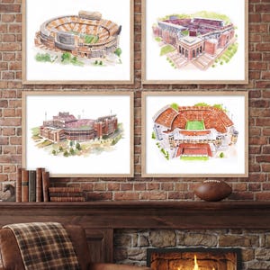 Pode incluir: Quatro impress&otilde;es em aguarela emolduradas com o t&iacute;tulo COLLEGIATE STADIUM PRINTS, cada uma representando um est&aacute;dio desportivo diferente em tons de laranja, vermelho e castanho. As molduras de madeira clara est&atilde;o numa parede de tijolo.
