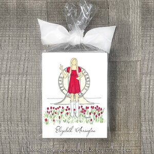 May include: A set of notecards featuring a hand-drawn illustration of a woman in a red dress standing in front of the University of Texas at Austin seal. The notecards are packaged in a cellophane bag with a ribbon and the name "Elizabeth Arrington" is printed below the illustration.