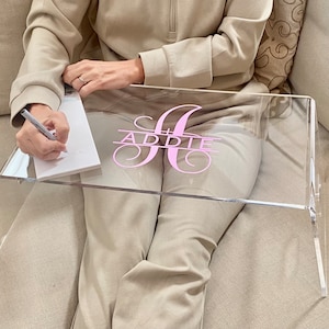 May include: A clear acrylic tray with a pink monogram that says "ADDIE" on it. The tray is sitting on a couch and a person is writing on a notepad on the tray.