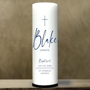 May include: A white pillar candle with a navy blue cross and script text that reads "Blake Louis." Below, the text "Baptized" is followed by the date "July 20, 2028" and "St. Charles Church."