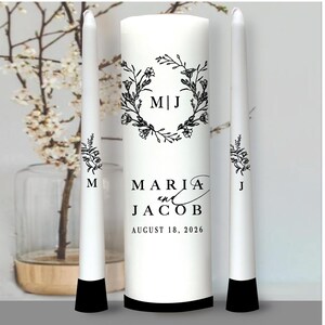 May include: Three white wedding candles with black bases. The center candle is taller and features a floral wreath with the initials "M|J" and the text "MARIA and JACOB AUGUST 18, 2026".
