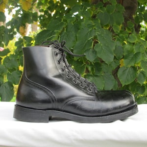 May include: A black leather boot with a lace-up design. The boot has a rounded toe and a sturdy sole. The laces are black, and the boot appears to be in good condition. The background is a blurred view of green foliage.