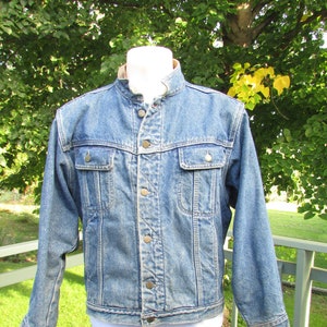 May include: A blue denim jacket with a stand-up collar and button closure. The jacket has two flap pockets on the front and a single pocket on the left chest.