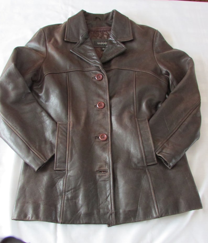 May include: A brown leather jacket with a button closure and a collar. The jacket has a label that says "SAHAR" on it.