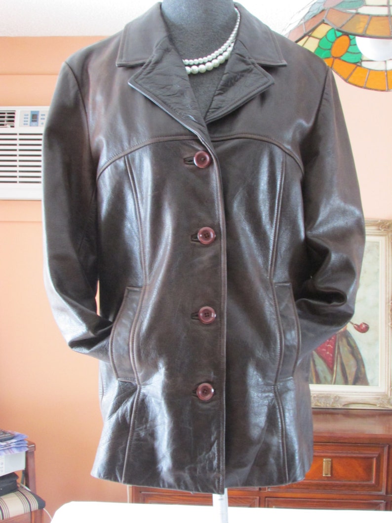 May include: A brown leather jacket with a button-up front and two pockets. The jacket has a collar and is lined with a brown fabric.