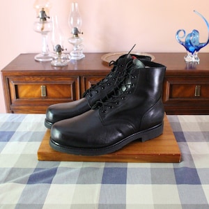May include: A pair of black leather lace-up boots with a thick sole, sitting on a wooden platform. The boots are on a checkered tablecloth.