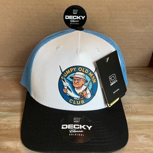 以下が含まれることがあります： 「Decky Classic Original」の野球帽。白いフロントパネルには、釣りをする老人の漫画が描かれています。帽子は黒いツバ、青いメッシュの背面、青い上端を備えています。「Grumpy Old Man Club」の文字が見えます。