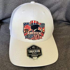 Peut inclure: Une casquette trucker blanche avec un dos en filet et un motif sur le devant repr&eacute;sentant une silhouette portant un haut-de-forme, des feux d'artifice, la Liberty Bell et le texte AMERICA 250. Un autocollant noir sur la visi&egrave;re indique TRUCKER.
