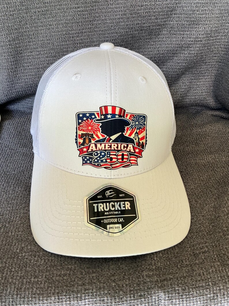 Peut inclure: Une casquette trucker blanche avec un dos en filet et un motif sur le devant repr&eacute;sentant une silhouette portant un haut-de-forme, des feux d'artifice, la Liberty Bell et le texte AMERICA 250. Un autocollant noir sur la visi&egrave;re indique TRUCKER.