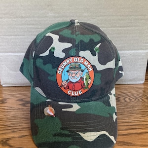 May include: Camouflage baseball cap with a "Grumpy Old Man Club" patch. The patch features a cartoon of an elderly man fishing, with the text "Grumpy Old Man Club" in a circular design. The cap is green, black, and white.