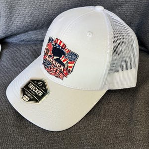 Peut inclure: Une casquette trucker blanche avec un arri&egrave;re en filet et un motif sur le devant repr&eacute;sentant l'Oncle Sam, des feux d'artifice et le texte AMERICA 250. Un autocollant noir et or sur la visi&egrave;re indique TRUCKER, EST. 1977, ADJUSTABLE, OUTDOOR CAP, ONE SIZE.