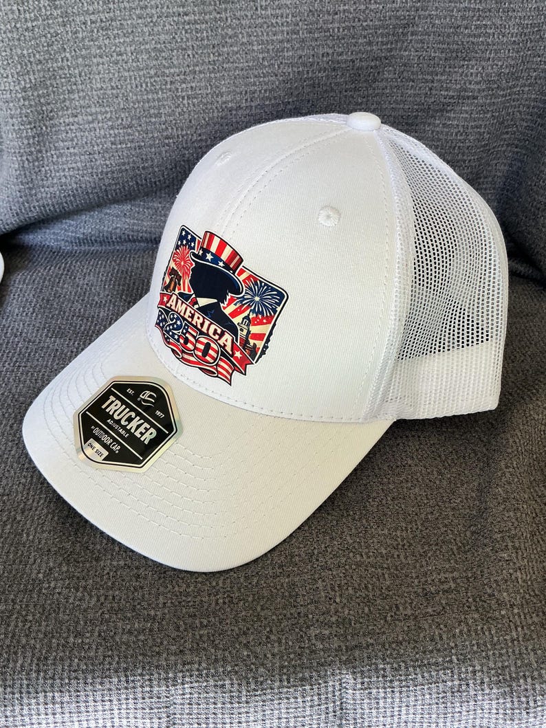 Peut inclure: Une casquette trucker blanche avec un arri&egrave;re en filet et un motif sur le devant repr&eacute;sentant l'Oncle Sam, des feux d'artifice et le texte AMERICA 250. Un autocollant noir et or sur la visi&egrave;re indique TRUCKER, EST. 1977, ADJUSTABLE, OUTDOOR CAP, ONE SIZE.