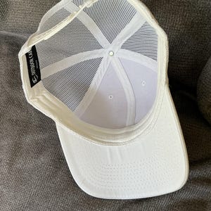Peut inclure: Une casquette de baseball blanche avec un panneau arri&egrave;re en maille et une fermeture snapback en plastique. L'int&eacute;rieur pr&eacute;sente une &eacute;tiquette en tissu noir avec le texte blanc OUTDOOR CAP. La casquette a un panneau avant blanc uni.