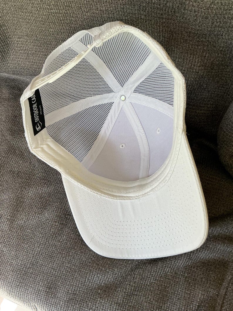 Peut inclure: Une casquette de baseball blanche avec un panneau arri&egrave;re en maille et une fermeture snapback en plastique. L'int&eacute;rieur pr&eacute;sente une &eacute;tiquette en tissu noir avec le texte blanc OUTDOOR CAP. La casquette a un panneau avant blanc uni.