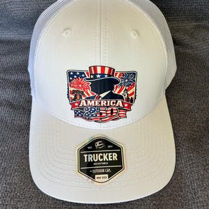 Peut inclure: Une casquette trucker blanche avec un dos en filet et un motif sur le devant repr&eacute;sentant l'Oncle Sam, la Liberty Bell, des feux d'artifice et le texte AMERICA 250. Un autocollant noir et argent sur la visi&egrave;re indique TRUCKER ADJUSTABLE BY OUTDOOR CAP ONE SIZE.