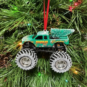 May include: A green monster truck Christmas ornament with the word "AVENGER" on the side. The truck has large black tyres with silver rims and a small green Christmas tree in the truck bed. The ornament hangs from a red ribbon.