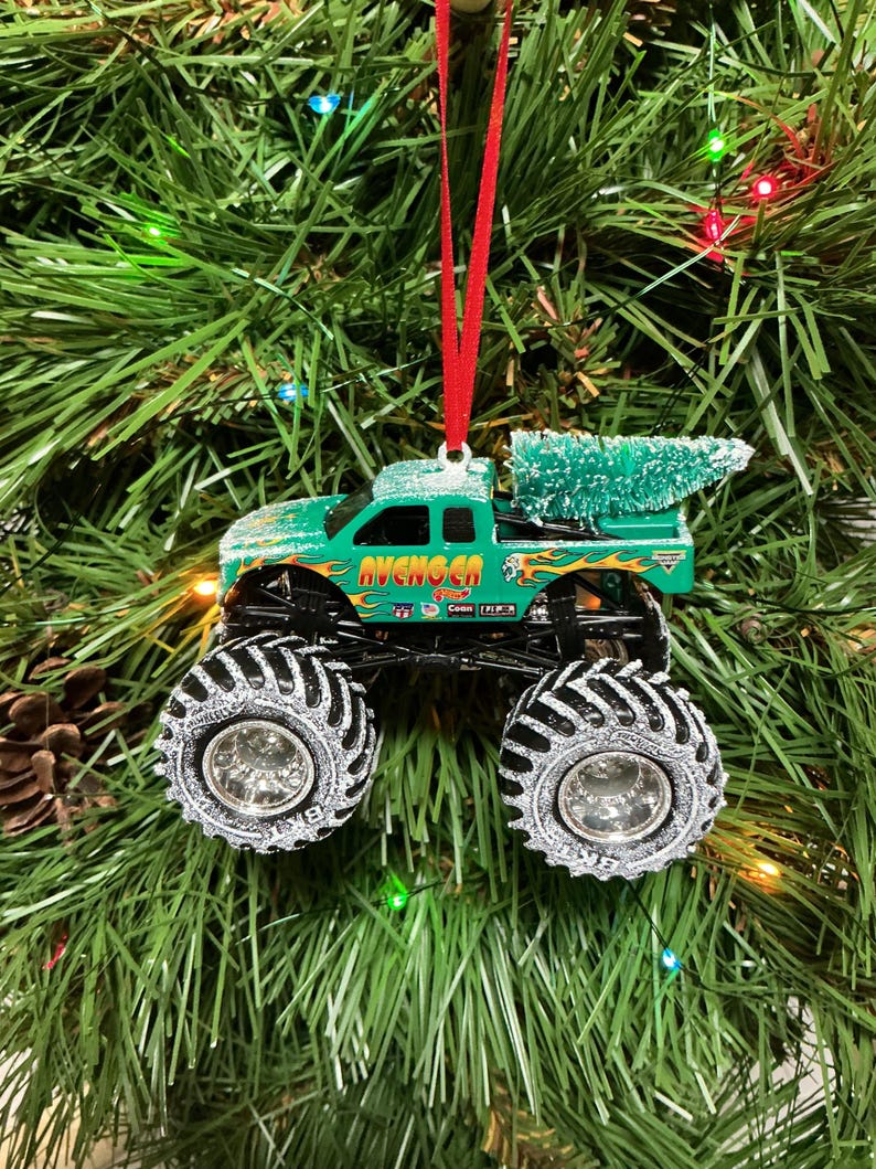 May include: A green monster truck Christmas ornament with the word "AVENGER" on the side. The truck has large black tyres with silver rims and a small green Christmas tree in the truck bed. The ornament hangs from a red ribbon.