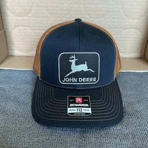 Pode incluir: Boné de caminhoneiro azul marinho e castanho com aba preta. O boné apresenta um patch retangular com um gráfico de veado branco e as palavras "JOHN DEERE". Há também uma etiqueta Richardson 112 Trucker.