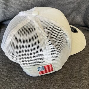 Peut inclure: Une casquette de baseball blanche avec un panneau arri&egrave;re en maille et un panneau avant solide. Un petit &eacute;cusson rectangulaire affichant le drapeau am&eacute;ricain est cousu sur la partie arri&egrave;re inf&eacute;rieure de la maille, avec une &eacute;tiquette noire.