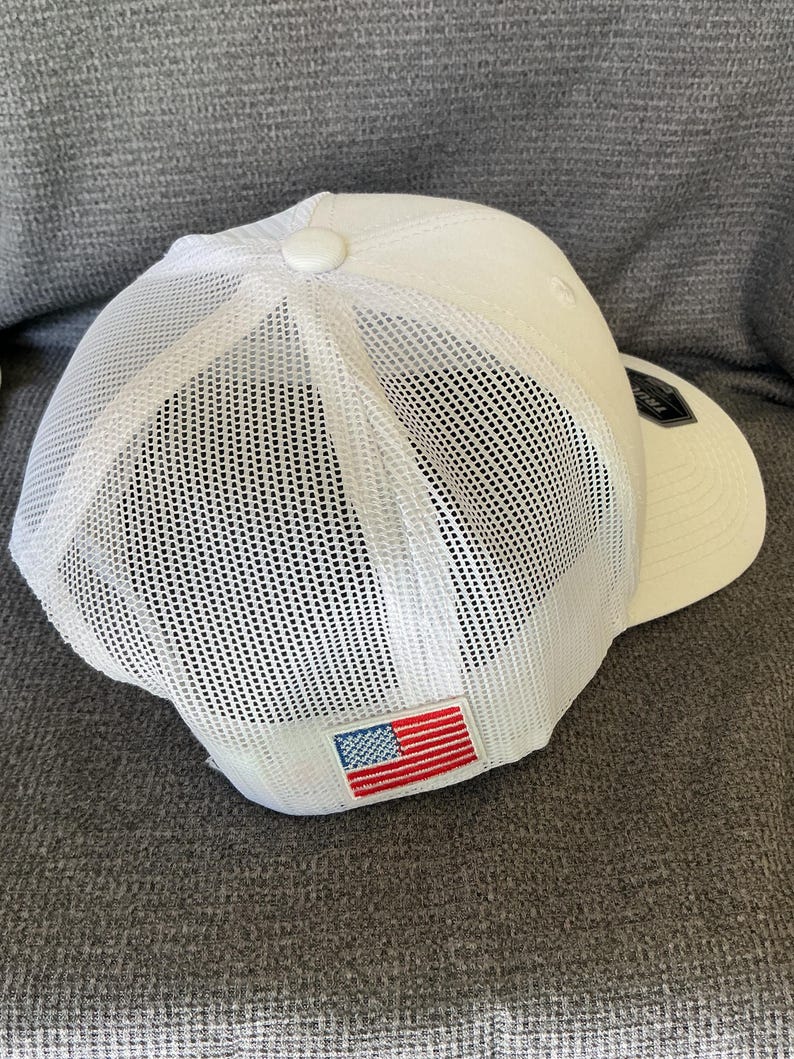 Peut inclure: Une casquette de baseball blanche avec un panneau arri&egrave;re en maille et un panneau avant solide. Un petit &eacute;cusson rectangulaire affichant le drapeau am&eacute;ricain est cousu sur la partie arri&egrave;re inf&eacute;rieure de la maille, avec une &eacute;tiquette noire.