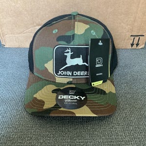 May include: Camouflage-patterned baseball cap with a black mesh back. The front features a rectangular patch with the John Deere logo and a white deer silhouette. A black tag with the Decky logo is attached.