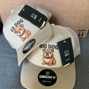 May include: Two tan baseball caps with a bulldog graphic and the text "LIL DOG" and "BIG DOG". The caps have a mesh back and a curved brim. Each cap has a black tag with the brand name "DECKY".