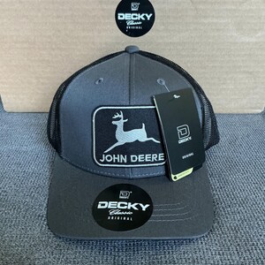 May include: A gray and black trucker hat with a mesh back and a curved brim. The hat features a rectangular patch with the John Deere logo and text. The hat also has a tag with the Decky logo.