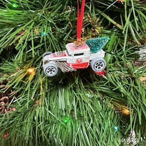 May include: A Christmas tree ornament shaped like a white and red toy truck with a small green tree in the truck bed. The ornament has silver wheels and is hanging from a red ribbon on a Christmas tree.
