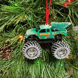 May include: Green monster truck Christmas ornament with the word "AVENGER" in orange and yellow flames. The truck has large black tyres with silver rims and a small green Christmas tree in the truck bed. The ornament hangs from a red ribbon.