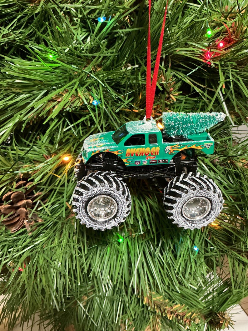 May include: Green monster truck Christmas ornament with the word "AVENGER" in orange and yellow flames. The truck has large black tyres with silver rims and a small green Christmas tree in the truck bed. The ornament hangs from a red ribbon.