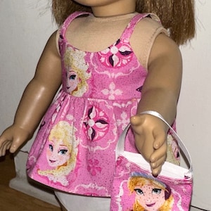 May include: A pink and white dress with a glittery pattern featuring a cartoon character. The dress has thin straps and a white underskirt. The doll is wearing a pink headband and holding a pink and white bag with a cartoon character.