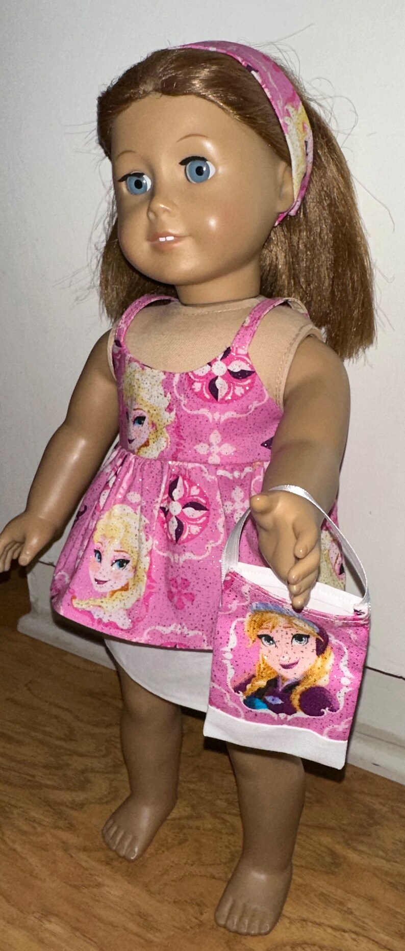 May include: A pink and white dress with a glittery pattern featuring a cartoon character. The dress has thin straps and a white underskirt. The doll is wearing a pink headband and holding a pink and white bag with a cartoon character.