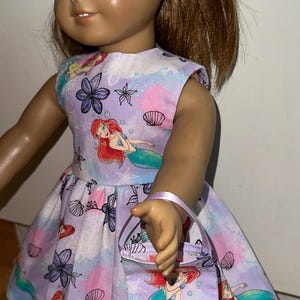 May include: A doll wearing a lavender dress, matching headband, and purse. The dress features a mermaid print with seashells and flowers. The doll has blue eyes and brown hair. The purse has a small handle.