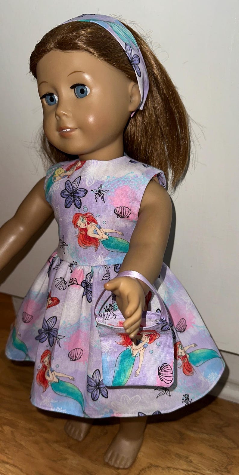 May include: A doll wearing a lavender dress, matching headband, and purse. The dress features a mermaid print with seashells and flowers. The doll has blue eyes and brown hair. The purse has a small handle.