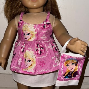 May include: A doll wearing a pink and white dress with a Frozen theme. The dress has a white underskirt and a pink bodice with images of Elsa and Anna from the movie Frozen. The doll is also wearing a headband with a pink and white pattern. The doll is holding a small white bag with a Frozen theme.