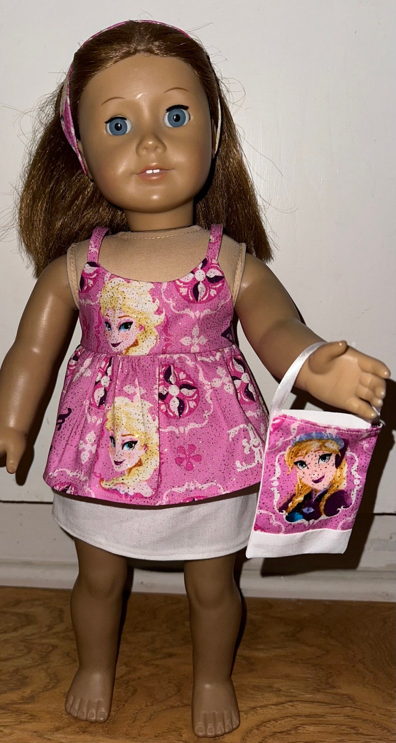 May include: A doll wearing a pink and white dress with a Frozen theme. The dress has a white underskirt and a pink bodice with images of Elsa and Anna from the movie Frozen. The doll is also wearing a headband with a pink and white pattern. The doll is holding a small white bag with a Frozen theme.